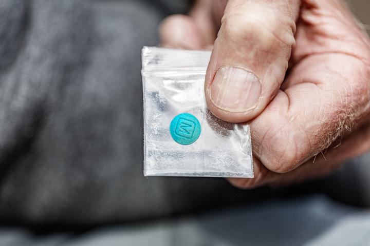 A hand holds a small plastic baggie with a blue pill