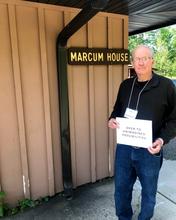 Bob Marcum stands in front of a building labeled "Marcum House" and he holds a sign reading "Open to unimagined possibilities."